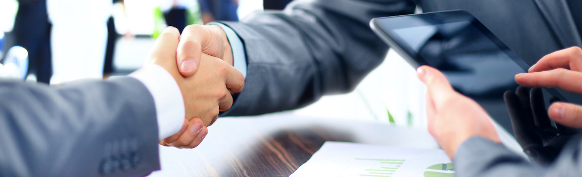 2 men in business attire shaking hands, man also working on an electronic tablet