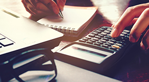 Hand using a calculator with laptop and notepad in the foreground