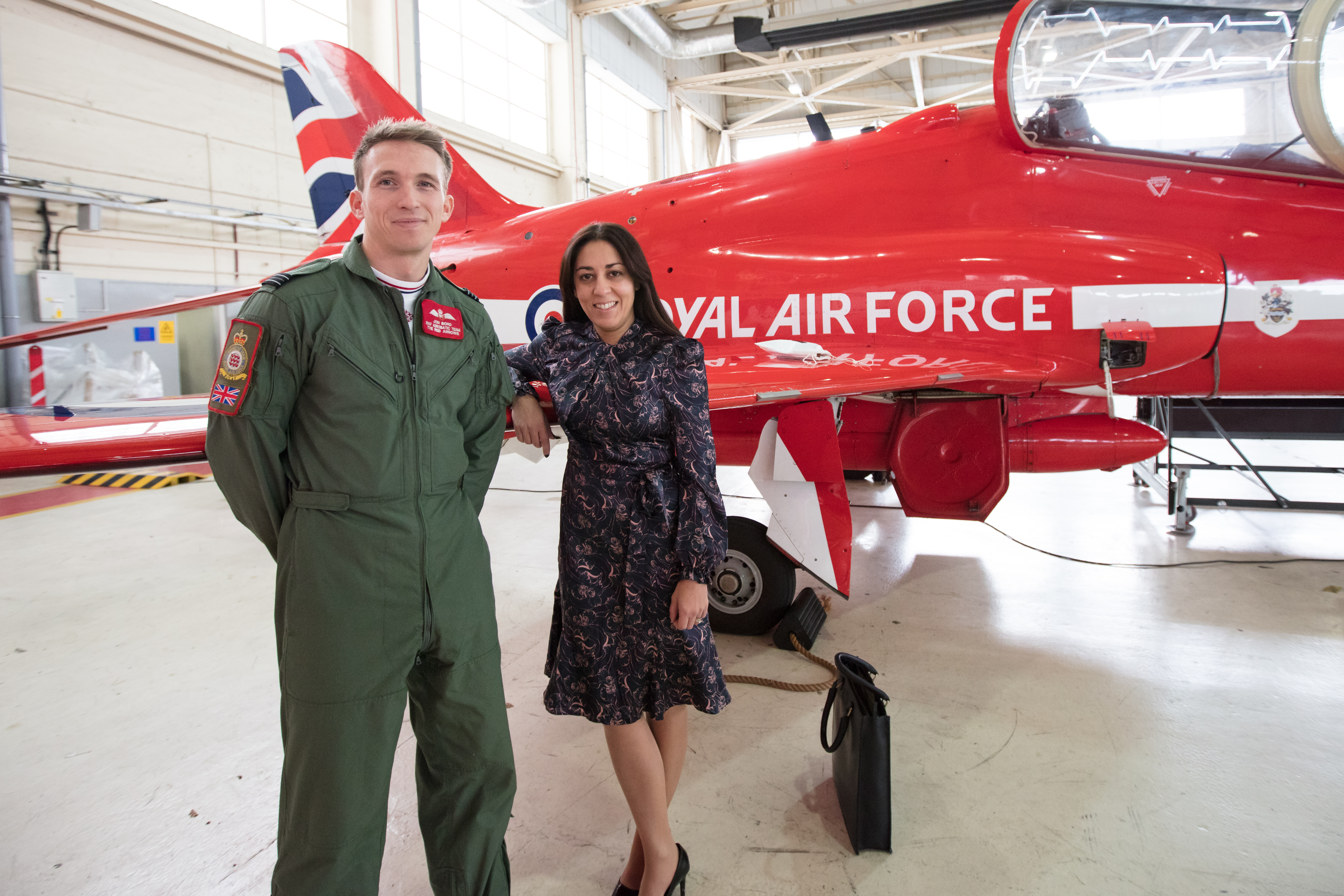 Lady stood next to gentleman in green flight suit with a RAF Red Arrow aeroplane in the background