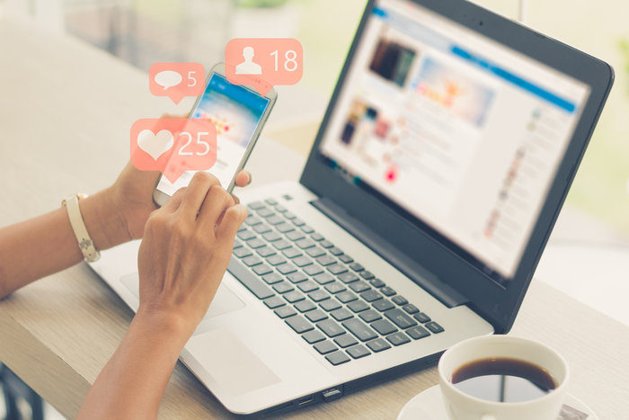 Lady using a social media app on a mobile phone with a laptop on the desk in front