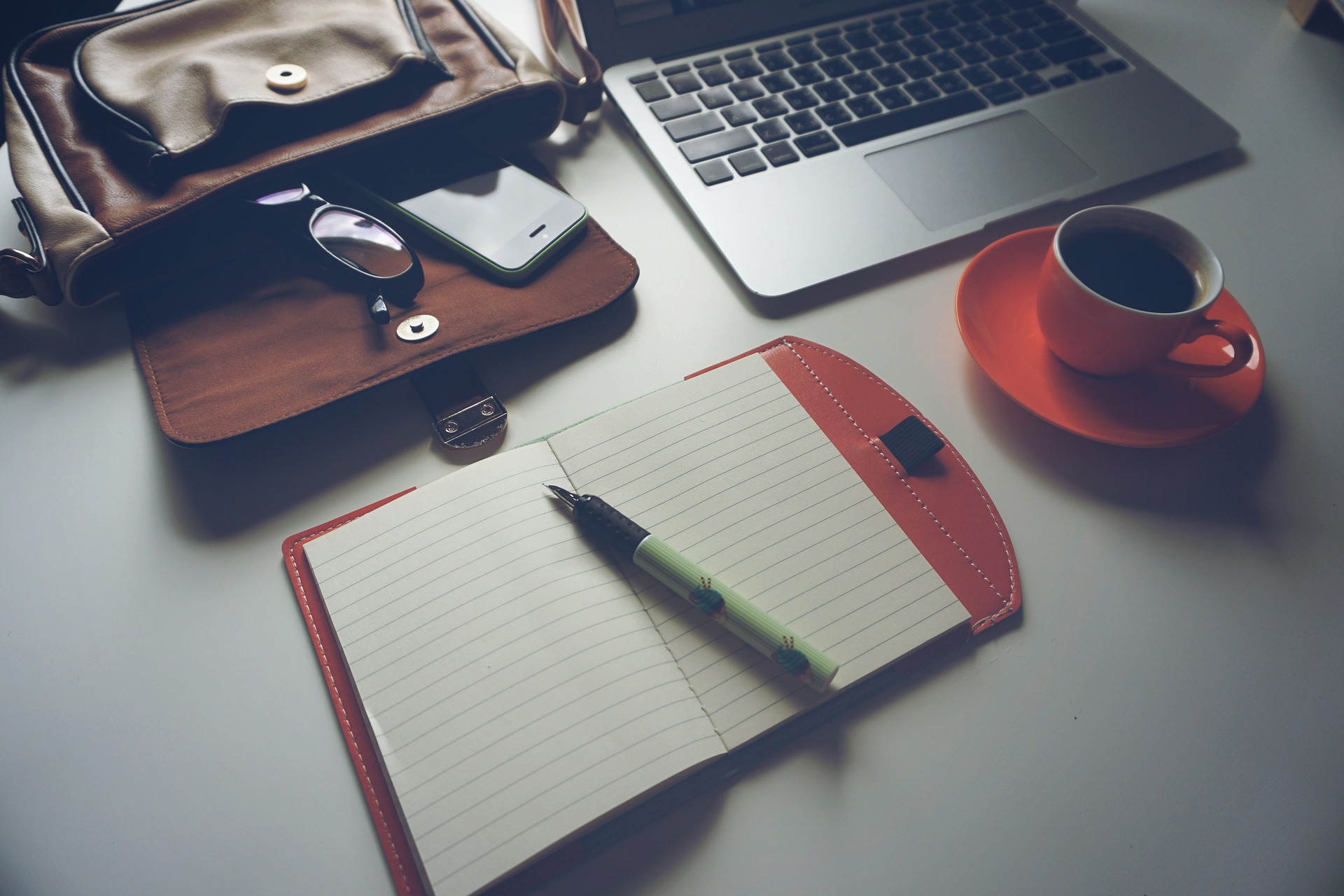 Open notepad with pen, cup of coffee, open laptop bag with glasses and phone, and laptop all on a desk