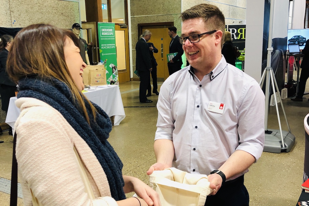 Gentleman holding a canvas bag open whilst talking to a lady at a trade show