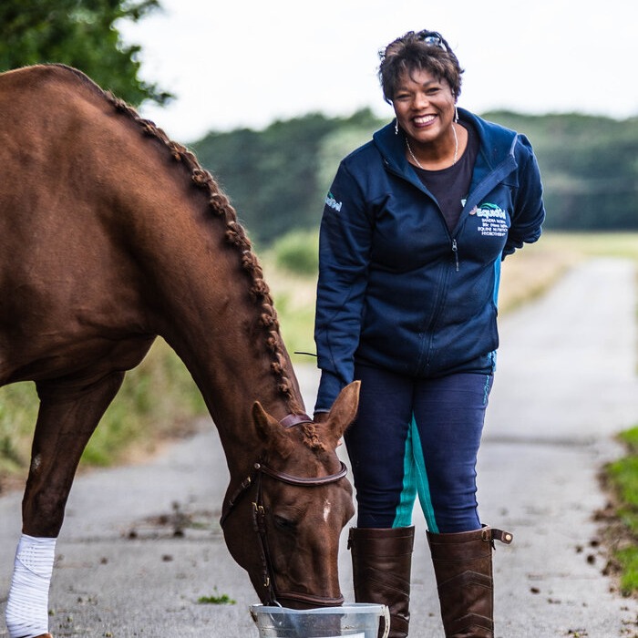 Lady stood next to a horse that is eating out of a bucket