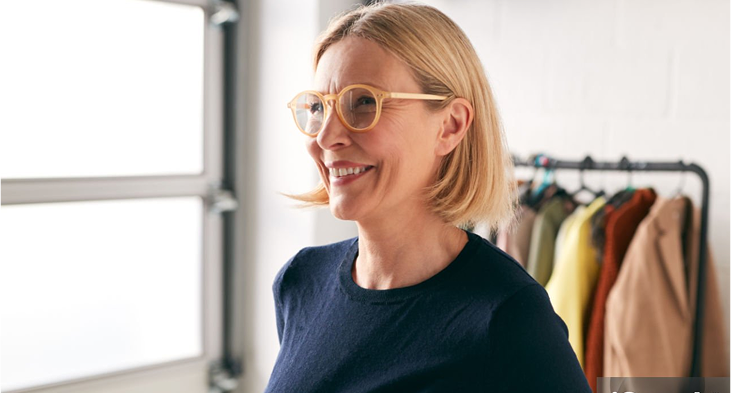 A woman with glasses and short blonde hair smiling with a rack of coats behind her