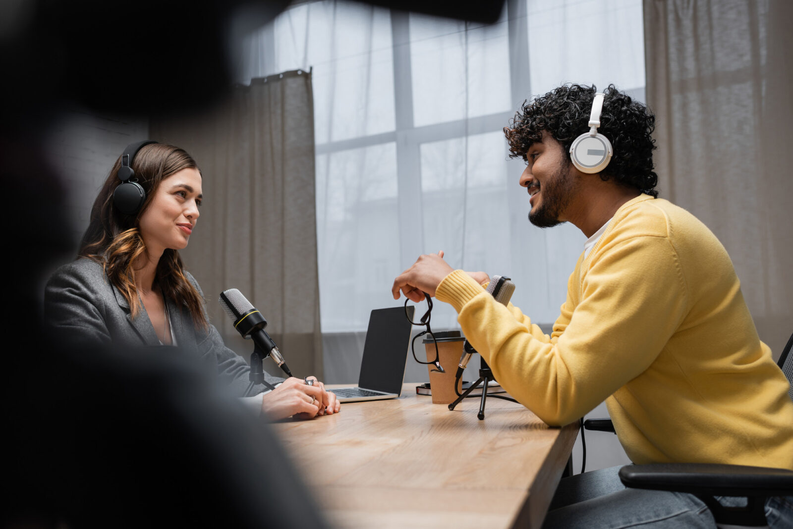 2 people with headphones on facing one another in an informal interview style chat, man holding glasses and lady sat in front of microphone with laptop next to her
