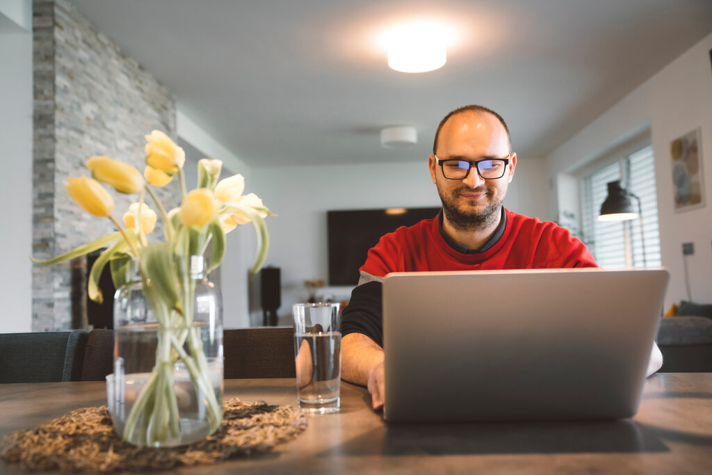 A man sat a desk at home with yellow flowers on the table, glass of water and typing at a desk - support and advice - Business Lincolnshire