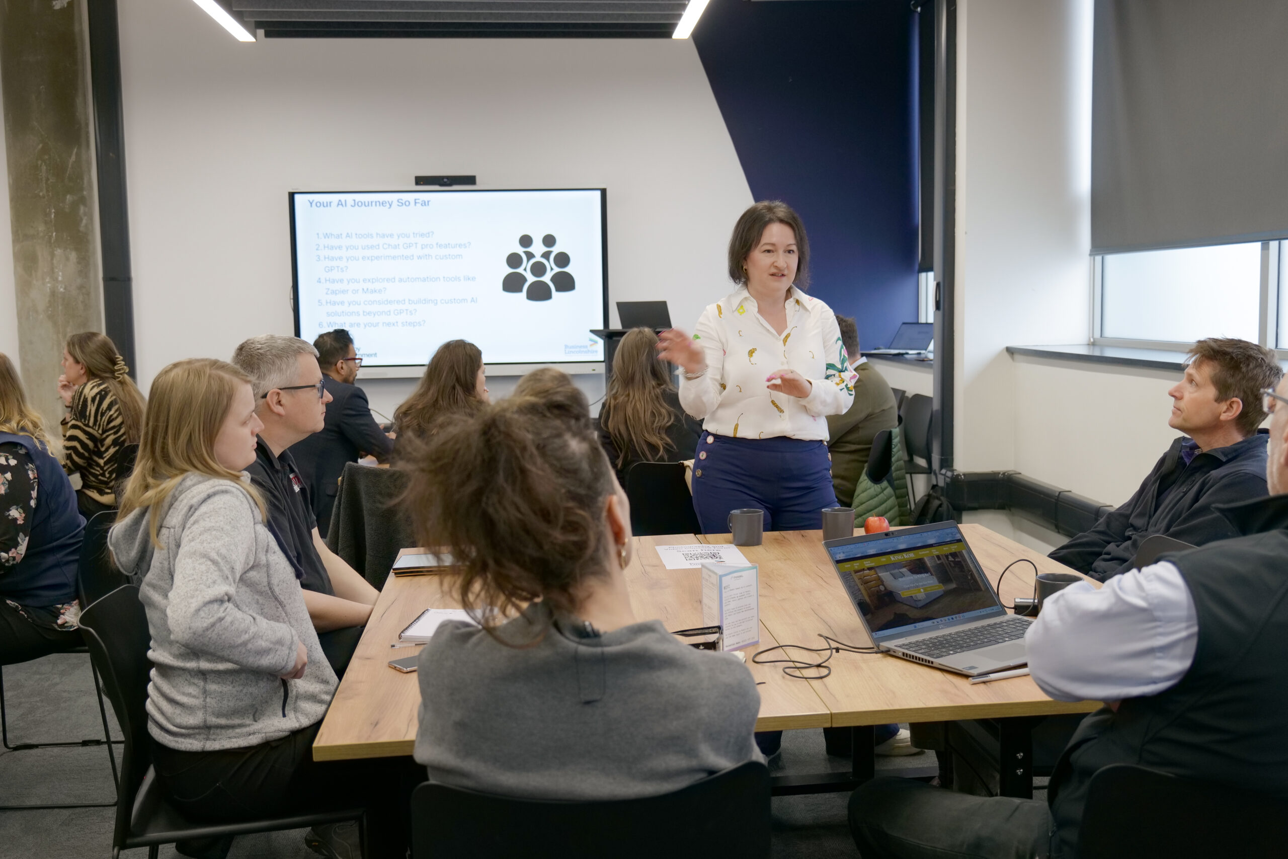 A group of people attending a presentation in a conference room. A presenter stands in front of a screen titled 'Your AI Journey So Far' with six bullet points about AI tools and usage. Attendees are seated around tables, some using laptops.